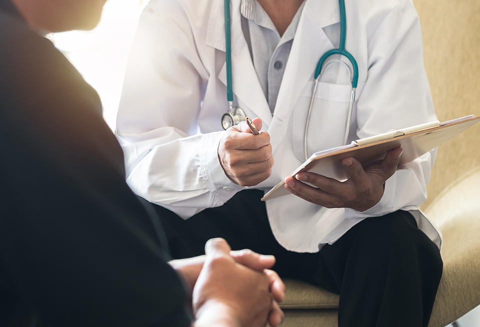 A doctor holding a clipboard and pen talks to a male patient.