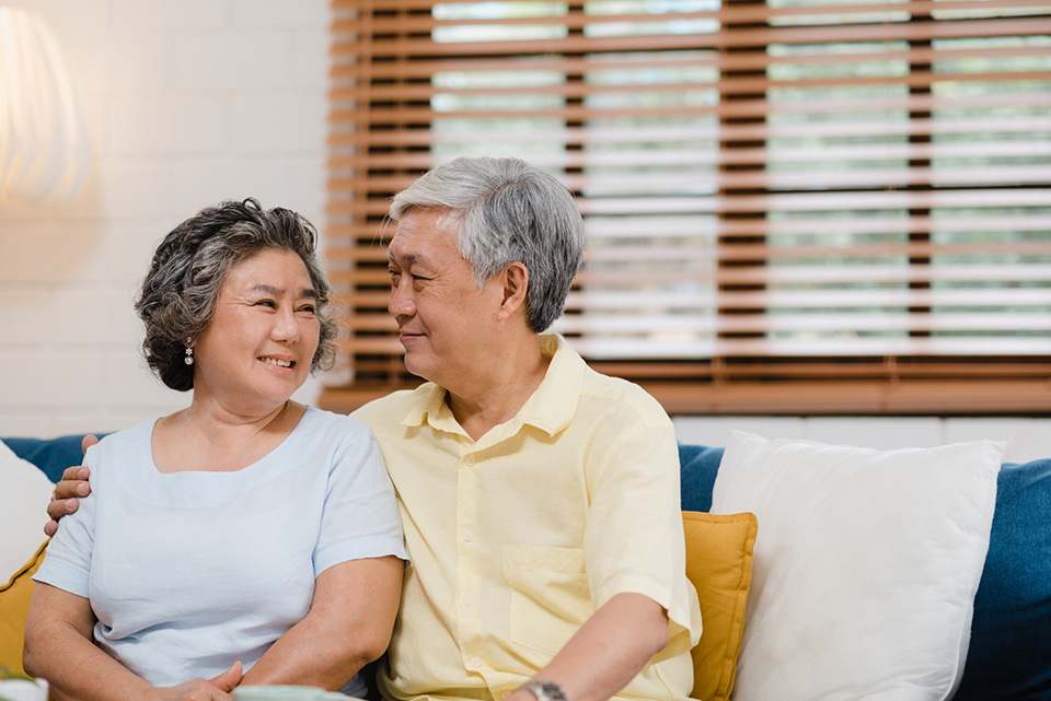 A woman and man sit on a couch, looking lovingly at each other. The man has his arm around the woman's shoulders.