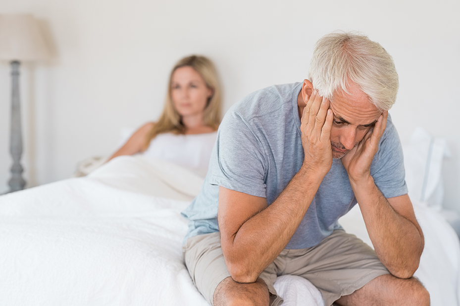 A man sits on the side of the bed with his head in his hands. His wife is in the background looking away from him.