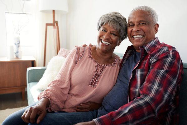 A woman and a man sit on a couch, smiling toward the camera. The woman leans her head against the man, who has an arm around her shoulders.