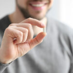 Closeup on the hand of a man holding a small white pill between his thumb and index finger.